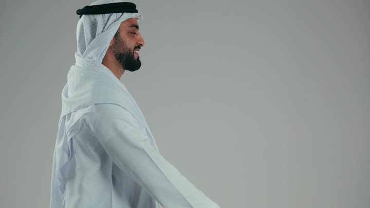 التسوق وشراء المستلزمات والهدايا عبر الانترنت، استخدام التكنولوجيا في الحياة اليومية، ايماءات وتعابير السرور، بورتريه من الجانب لشاب عربي خليجي اماراتي يرتدي الغترة والكندورة يمسك بيده حقيبه ورقيه حمراء، خلفية رماديه