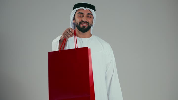 Smiling young Emirati Arab man wearing a white kandura and ghutra holding a red paper bag and offering it to someone, gestures of joy and happiness, the art of giving gifts, shopping and buying gifts, acquiring important things, gray background