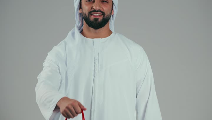 شاب عربي خليجي اماراتي مبتسم يرتدي الكندوره والغتره البيضاء يحمل كيس ورقي احمر ويقدمه لاحد ما, ايماءات الفرح والسرور , فن تقديم الهدايا, التسوق وشراء الهدايا, اقتناء الاشياء الهامه, خلفيه رماديه