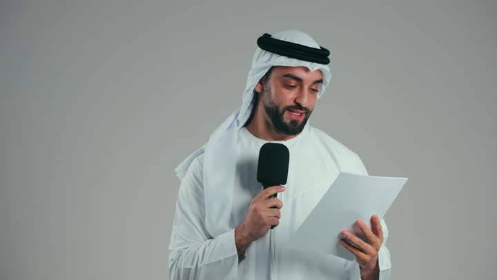 Exchanging dialogue, discussion and effective communication, giving speeches from the paper with gestures of self-confidence, presenting workshops and training courses, the art of speaking and public speaking, a portrait of a young Arab Gulf Emirati man wearing a kandura and a ghutra, standing confidently holding a loudspeaker, gray background