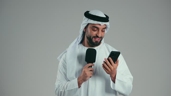 إقامة ورش العمل والدورات التدريبية، فن التحدث والخطابة، التواصل الفعال والنقاش، بورتريه لشاب عربي خليجي اماراتي يرتدي الكندورة والغترة يمسك مكبر الصوت يقوم بالقراءة من الهاتف المحمول، إلقاء الخطاب بإيماءات الثقة بالنفس، خلفية رماديه