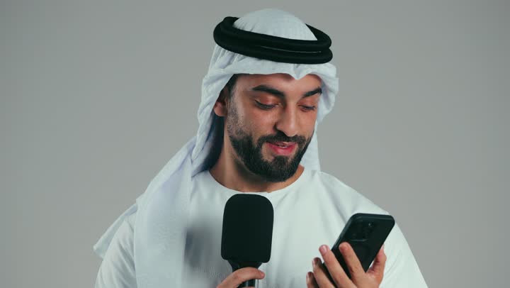 Holding workshops and training courses, the art of speaking and public speaking, effective communication and discussion, portrait of a young Arab Gulf Emirati man wearing a kandura and ghutra holding a loudspeaker reading from a mobile phone, delivering a speech with self-confident gestures, gray background