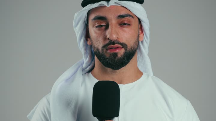 Giving lectures and holding seminars, exchanging dialogue and discussion and effective communication, presenting workshops and training courses, the art of speaking and public speaking, portrait of a young Emirati Gulf Arab man wearing a kandura and ghutra, standing confidently holding a loudspeaker, gray background