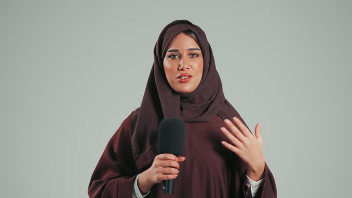 Speaking and dialogue skills, gestures and expressions of integration, focus and self-confidence, an Arab Gulf Emirati woman presenting a segment, a Saudi woman wearing a hijab and an abaya looking at the camera holding a microphone, mastery of speaking and delivery, gray background