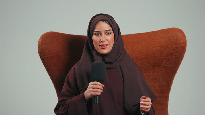 Speaking and dialogue skills, mastering the art of public speaking and oratory, a Saudi woman sitting on a chair presenting workshops and training courses using a loudspeaker, a side portrait of a veiled young Arab Gulf Emirati woman wearing an abaya holding the microphone with gestures of integration and focus, white background