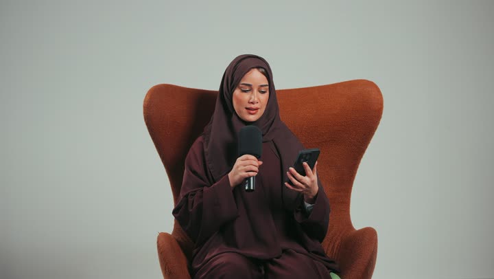 إقامة ورش العمل والدورات التدريبية، التواصل الفعال وتبادل الحوار والنقاش، إمرأة سعودية تجلس على الكرسي تقوم بالقراءة من الهاتف المحمول، بورتريه لشابة عربية خليجية اماراتية محجبة ترتدي العباءة تمسك مكبر الصوت تلقي الخطابات، خلفية رماديه