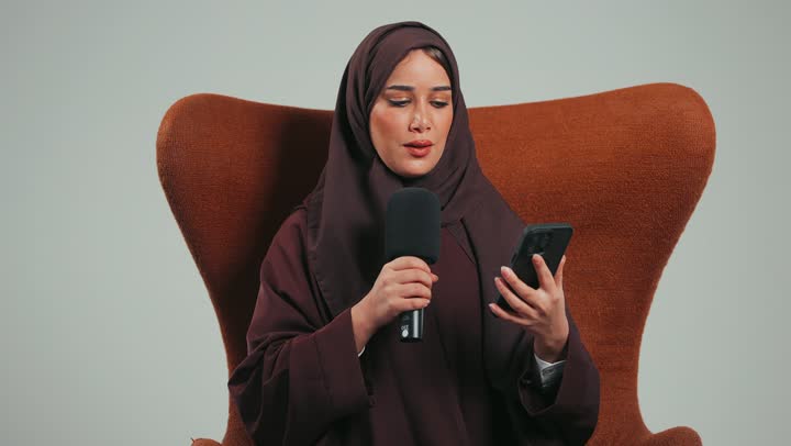 Holding workshops and training courses, effective communication and exchange of dialogue and discussion, a Saudi woman sitting on a chair reading from a mobile phone, a portrait of a veiled young Arab Gulf Emirati woman wearing an abaya holding a loudspeaker giving speeches, gray background