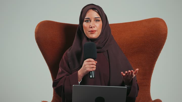 Saudi woman sitting on a chair reading from a laptop, holding workshops and training courses, portrait of a veiled young Arab Gulf Emirati woman wearing an abaya holding a microphone delivering a speech with confident gestures, the art of speaking and public speaking, gray background