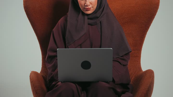 سيده اماراتيه تستخدم الأجهزة والتقنيات المتطورة, بورتريه لإمرأة عربية خليجية سعودية ترتدي العباءة تعمل على الحاسوب, إدارة وإنجاز الأعمال عبر الاونلاين, التسوق الالكتروني عبر التطبيقات والتكنولوجيا الحديثة, إجراء مقابلة عمل عن بعد, خلفية رماديه