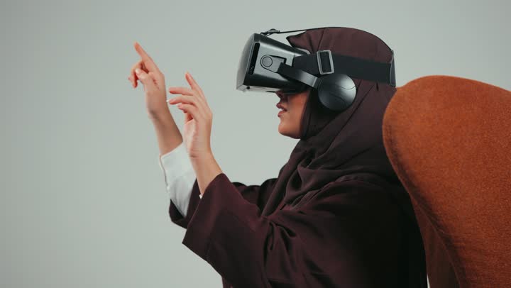 Virtual reality glasses, Saudi woman clicking on something, using modern technical devices, portrait of an Arab Gulf Emirati woman wearing hijab and abaya sitting on a chair wearing RV glasses, simulating the imaginary world, gray background