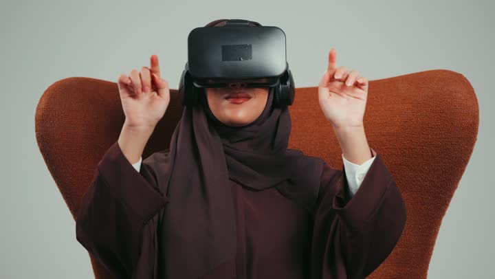 محاولة لمس شيء ما، تعابير الاندماج والتفاعل، بورتريه لإمرأة عربية خليجية اماراتية ترتدي الحجاب والعباءة ونظارات الواقع الافتراضي تجلس على الكرسي تقوم بمد يديها الى الامام ومحاكاة العالم الخيالي، شابة سعودية تستخدم نظارة الفي آر، خلفية رماديه