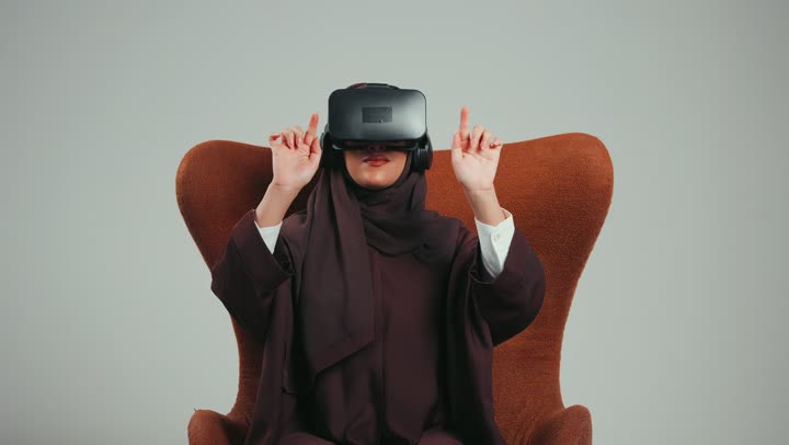 محاولة لمس شيء ما، تعابير الاندماج والتفاعل، بورتريه لإمرأة عربية خليجية اماراتية ترتدي الحجاب والعباءة ونظارات الواقع الافتراضي تجلس على الكرسي تقوم بمد يديها الى الامام ومحاكاة العالم الخيالي، شابة سعودية تستخدم نظارة الفي آر، خلفية رماديه