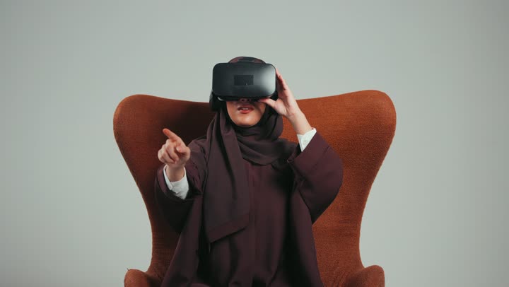 محاكاة العالم الخيالي والاندماج والتفاعل، بورتريه لإمرأة عربية خليجية اماراتية ترتدي الحجاب والعباءة ونظارات الواقع الافتراضي تجلس على الكرسي تقوم بمد يديها الى الامام بتعابيرالنقرعلي شيء ما، شابة سعودية تستخدم نظارة الفي آر، خلفية رماديه