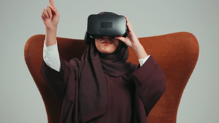 محاكاة العالم الخيالي والاندماج والتفاعل، بورتريه لإمرأة عربية خليجية اماراتية ترتدي الحجاب والعباءة ونظارات الواقع الافتراضي تجلس على الكرسي تقوم بمد يديها الى الامام بتعابيرالنقرعلي شيء ما، شابة سعودية تستخدم نظارة الفي آر، خلفية رماديه