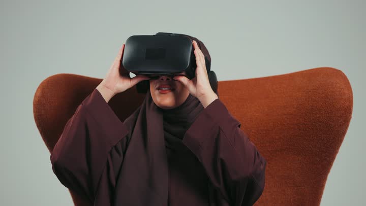 التقنيات الحديثة والمتطوره، مفهوم التفاعل والاندماج، محاكاة العالم الخيالي، بورتريه لامرأه عربية خليجية سعودية محجبة ترتدي عباءه ملونه تضع نظارة الفي ار ثلاثية الأبعاد، سيده اماراتيه تجلس علي كرسي تمسك بيدها نظاره الابعاد الثلاثيه،  خلفية رماديه