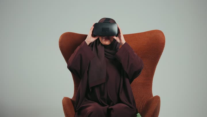 التقنيات الحديثة والمتطوره، مفهوم التفاعل والاندماج، محاكاة العالم الخيالي، بورتريه لامرأه عربية خليجية سعودية محجبة ترتدي عباءه ملونه تضع نظارة الفي ار ثلاثية الأبعاد، سيده اماراتيه تجلس علي كرسي تمسك بيدها نظاره الابعاد الثلاثيه،  خلفية رماديه