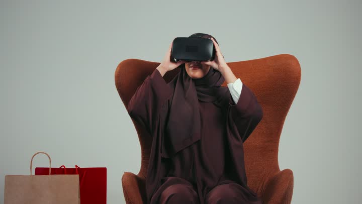 دمج التقنيات الحديثة بالتسوق، مفهوم التفاعل والاندماج، محاكاة العالم الخيالي، بورتريه لامرأه عربية خليجية سعودية محجبة ترتدي عباءه ملونه تضع نظارة الفي ار ثلاثية الأبعاد، سيده اماراتيه تجلس علي كرسي بجانب أكياس التسوق،  خلفية رماديه