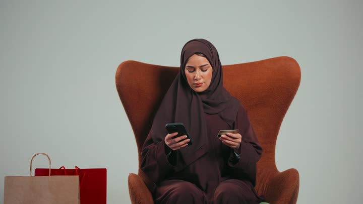 الاستفادة من الخصومات على المنتجات، الاستمتاع بالتسوق، شراء المستلزمات والاحتياجات الضرورية، بورتريه لسيده عربيه خليجيه اماراتيه تجلس علي كرسي تستخدم الهاتف للدفع الالكتروني، إمراه سعوديه تستمتع بالتسوق الالكتروني، خلفية رماديه