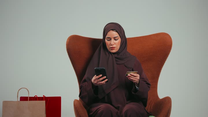 الاستفادة من الخصومات على المنتجات، الاستمتاع بالتسوق، شراء المستلزمات والاحتياجات الضرورية، بورتريه لسيده عربيه خليجيه اماراتيه تجلس علي كرسي تستخدم الهاتف للدفع الالكتروني، إمراه سعوديه تستمتع بالتسوق الالكتروني، خلفية رماديه