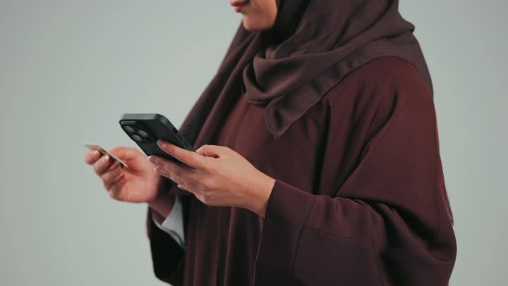الشراء والدفع عن طريق بطاقة الإئتمان البنكية، إيماءات وتعابير السعادة والسرور، إمكانية تسديد الفواتير بالبطاقات الإلكترونية، بورتريه من الجانب لسيدة عربية خليجية سعودية محجبة تحمل بيدها بطاقة الصراف، إمراه اماراتيه تتسوق عبر الانترنت، خلفية رمادية