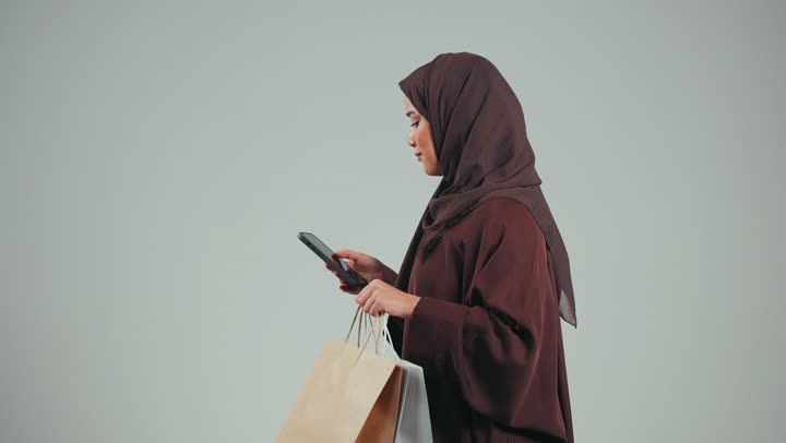 Using a bank credit card, taking advantage of electronic payment services, enjoying online shopping, side portrait of a Saudi Arabian Gulf woman wearing a colorful abaya, an Emirati woman using a phone for online orders, facilitating buying and selling operations, gray background