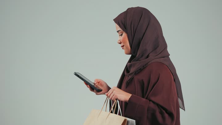 استخدام بطاقة الائتمان البنكية، الاستفادة من خدمات الدفع الالكتروني، الاستمتاع بالتسوق عبر الانترنت، بورتريه من الجانب لسيده عربية خليجية سعودية ترتدي عباءه ملونه، إمراءه امراتيه تستخدم الهاتف لطلبات الاون لاين، تسهيل عمليات الشراء والبيع ، خلفية رماديه
