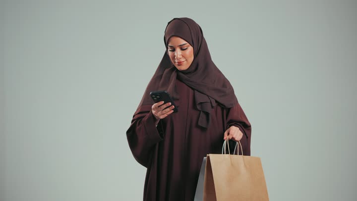 إمرأة سعودية تقوم بالتسوق وشراء المستلزمات, بورتريه من الجانب لسيدة عربية خليجية اماراتية مبتمسة  ترتدي الحجاب والعباءة البنية تعطي أكياس كرتون ملونه لشخص ما، ايماءات السرور والسعادة بتقديم الهدية، الاستفادة من العروض والخصومات، خلفية رماديه