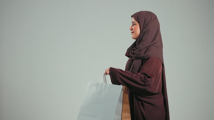 تعابير وايماءات السرور والسعادة بتقديم الهدية، الاستفادة من العروض والخصومات، إمرأة سعودية تقوم بالتسوق وشراء المستلزمات, بورتريه لسيدة عربية خليجية اماراتية مبتمسة ترتدي الحجاب والعباءة البنية تحمل بيدها أكياس كرتون ملونه، خلفية بيضاء