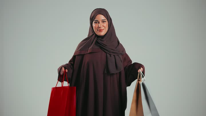 الاثر الايجابي للتسوق على نفسية النساء، الاستفادة من العروض والخصومات,  بورتريه لإمرأة عربية خليجية سعودية محجبة ترتدي العباءة تحمل بيدها أكياس كرتون ملونه تنظر الى الكاميرا بإيماءات الفرح والسرور, سيده اماراتيه مبتسمه تقدم الهدايا، خلفية رمادية