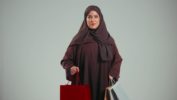 بورتريه لإمرأة عربيه خليجية سعوديه ترتدي عباءه ملونه أنيقه تحمل بيدها أكياس التسوق, التسوق الإلكتروني, ماركات عالمية, التسوق في المراكز التجاريه, متعة التسوق في السعودية,سيده اماراتيه تقدم حقيبه حمراء, عروض وتخفيضات موسميه, خلفيه رماديه