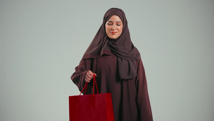 Offers and discounts on all items, seasonal discounts and sales, portrait of a veiled Saudi Arabian Gulf woman wearing an abaya, holding a red paper bag, looking at the camera with gestures of pleasure and happiness, an Emirati woman buying supplies and gifts, gray background