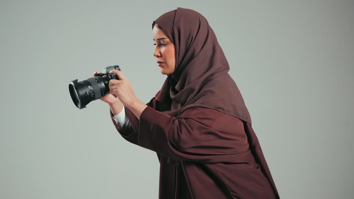 موهبه التصوير الفوتوغرافي عند النساء، ايماءات التركيزعند التقاط الصور، سيده سعوديه ترتدي الحجاب والعباءه الملونه تقوم باستخدام كاميرا رقميه عاليه الدقه، إمرأه اماراتيه تحترف مهنه التصوير، خلفيه رماديه