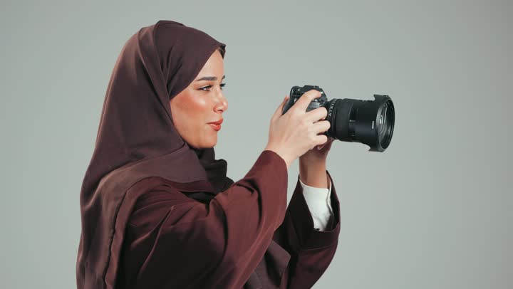 مصورة محترفة عربية سعودية خليجية تقوم بالتقاط صور فوتوغرافية, وظيفة ومهنة نسائية, بورتريه لإمراة اماراتية ترتدي الحجاب والعباءة تقوم بالتقاط صورعبر كاميرا فوتوغرافية، معدات وتقنيات متطورة، خلفية رماديه