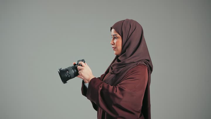 احتراف فن التصوير الفوتوغرافي باستخدام كاميرات رقمية عالية الدقة, وظيفة ومهنة نسائية, بورتريه لإمراة عربية خليجية اماراتية ترتدي الحجاب والعباءة تقوم بالتقاط صورة تذكارية عبر الكاميرا, مصورة سعودية تمارس هواية التصوير بتعابير السرور، خلفية رماديه
