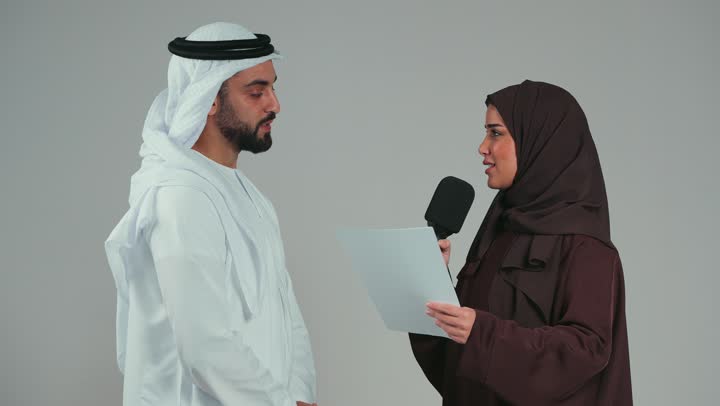 An Arab Emirati Gulf woman wearing a colorful hijab and abaya holding a bunch of white papers and a loudspeaker discussing with a man wearing a white kandura and ghutra. Effective communication and exchanging questions. The concept of dialogue and exchanging opinions. Gray background.