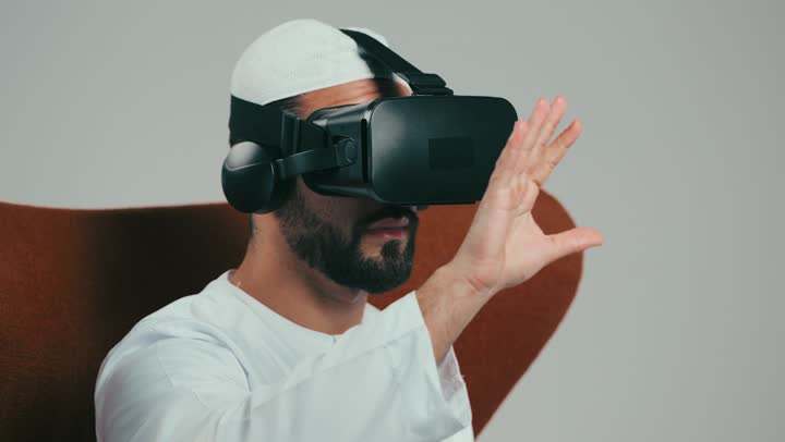 Combining modern technology and technology with fun and entertainment, simulating the imaginary world, portrait of an Emirati Gulf Arab man wearing a kandura and 3D virtual reality glasses sitting on a brown chair, gestures of awe, fear and repelling something, gray background