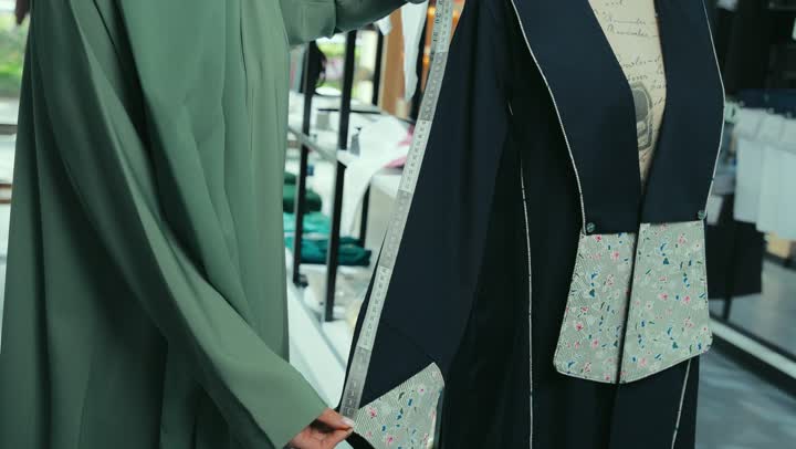 A Saudi woman measuring the length of the sleeve, buying new clothes and accessories, a photo of an Arab Gulf Emirati woman wearing an abaya taking the abaya size using a measuring tape, the profession of sewing and tailoring modern clothes, fashion show and new models