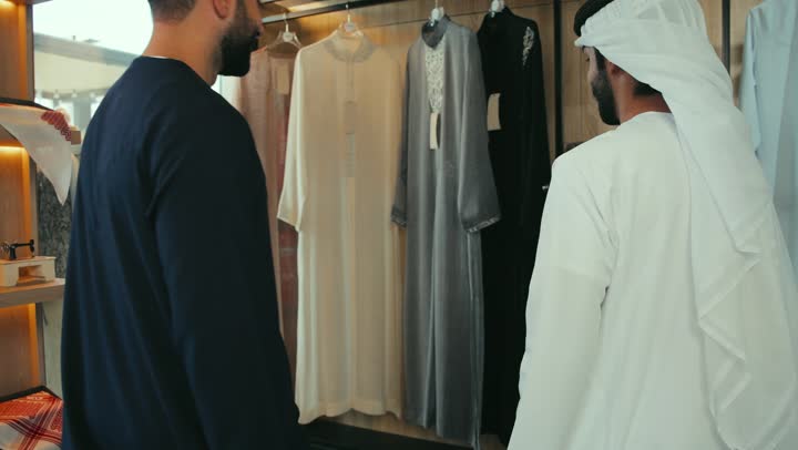 إيماءات السعادة و السرور،الاستفادة من العرض و الخصم الموسمي, تصوير مقرب لزبون عربي خليجي اماراتي مبتسم يرتدي كندورة كحلي يقوم باختيار ثوب من متجر من متجر المستلزمات الرجاليه , الاستمتاع بالتسوق في المول التجاري في الامارات العربيه المتحده