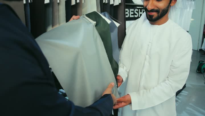Shopping in a Gulf clothing store, choosing the best fabrics for making clothes, sewing skills and mastery of tailoring youth clothes, a men's tailoring and tailoring shop, taking advantage of offers and discounts, a photo of a young Emirati Gulf Arab man wearing a navy blue kandura working as a tailor, showing it to a customer.