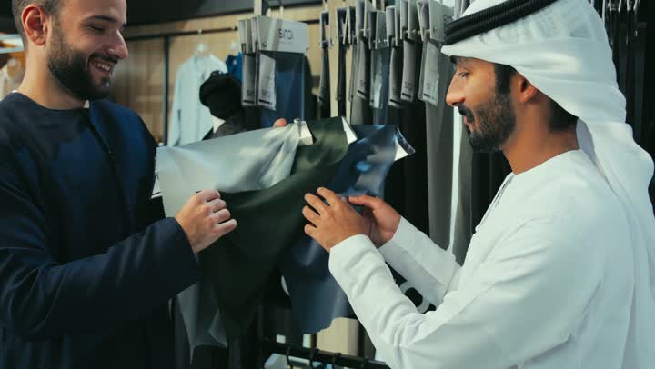 محل خياطة و تفصيل الملابس الرجالي ، الاستفاده من العروض والخصومات ،التسوق داخل متجر لوازم الزي الخليجي ،مهارة الخياطة واتقان تفصيل الملابس الشبابية ، تصوير لشاب عربي خليجي اماراتي يرتدي الكندورة الكحلي يعمل في مهنة الخياطة يعرض للزبون ، اختيار أجود أنواع الأقمشة لصنع الثياب .