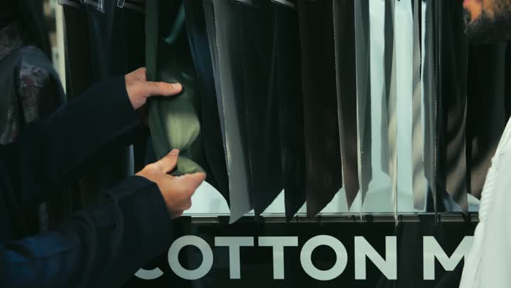 A photo of a young Emirati Gulf Arab man wearing a navy blue kandura working as a tailor showing customers how to benefit from offers and discounts, the best types of kanduras, choosing the best fabrics for making clothes, a men's tailoring and tailoring shop, sewing skills and mastery of tailoring youth clothes.