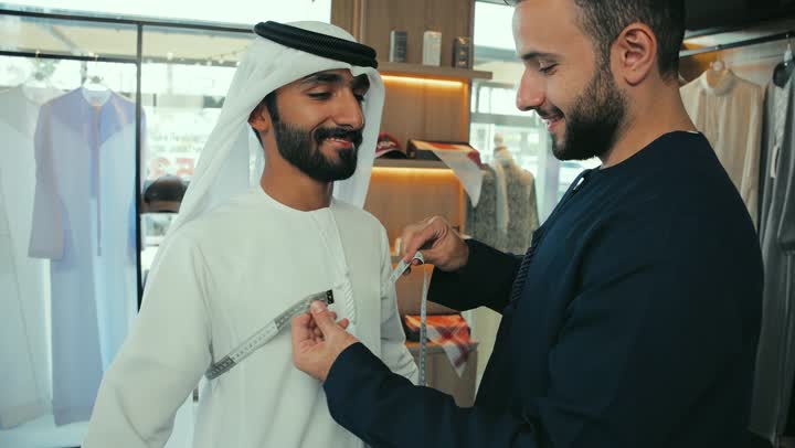 Men's tailoring and tailoring shop, tailoring, handicrafts, close-up of a young Emirati man wearing a navy blue kandura taking measurements of a customer's body using a tape measure, sewing skill and mastery of tailoring youth clothing