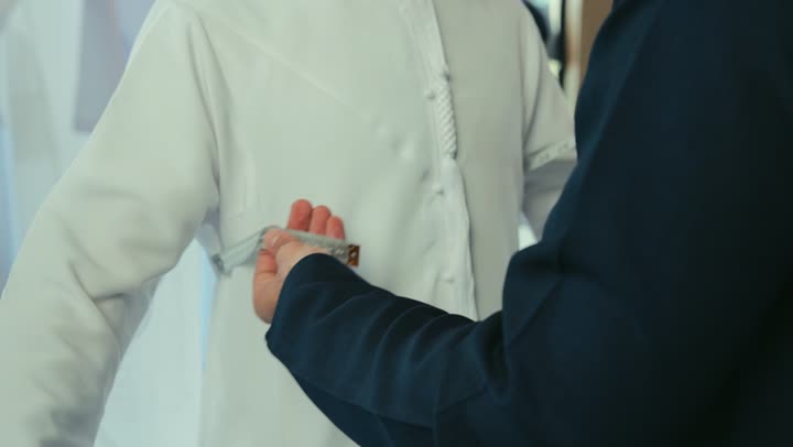Practicing the tailoring profession, handicrafts and crafts, a close-up of a young Emirati Gulf man wearing a navy blue kandura taking measurements of a customer's body using a tape measure, a men's tailoring and tailoring shop, sewing skills and mastery of tailoring youth clothing