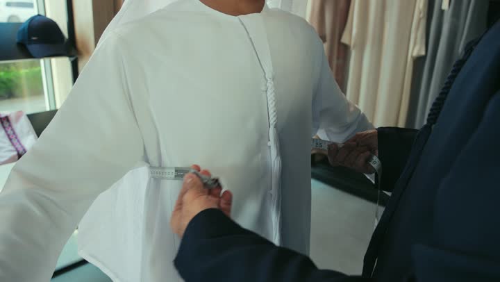 مفهوم عالم الازياء والموضه ، تصوير مقرب لشاب خليجي اماراتي يرتدي الكندورة الكحلي يقوم بأخذ القياسات الخاصة بجسم الزبون باستخدام الشريط المتري ، حرف ومهن يدوية ، مهارة اتقان تفصيل الملابس الشبابية، محل خياطة و تفصيل الملابس الرجالي ، اتقان صنع الملابس، ممارسة مهنة الخياطة.