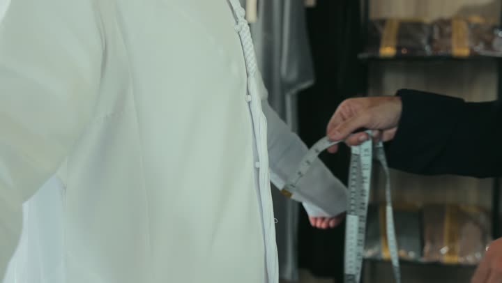 Close-up of a young Emirati man wearing a navy blue kandura taking measurements of a customer's body using a tape measure. Handicrafts and crafts, skill of mastering tailoring youth clothing, men's tailoring and tailoring shop, concept of the world of fashion and style, mastering clothing making, practicing the tailoring profession.