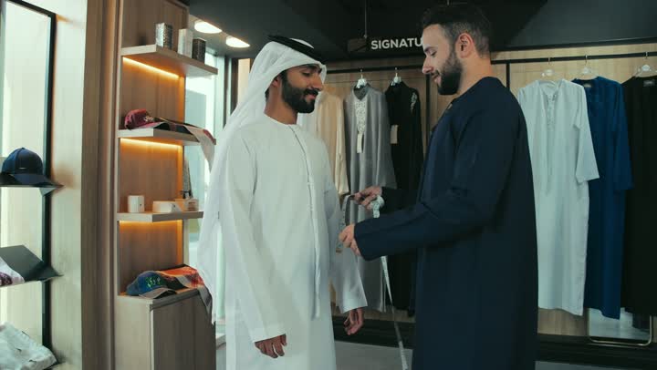 Handicrafts and trades, skill of mastering tailoring youth clothes, men's tailoring and tailoring shop, concept of the world of fashion and style, mastering clothing making, practicing the tailoring profession, close-up of a young Emirati Gulf man wearing a navy blue kandura taking measurements of a customer's body using a measuring tape.