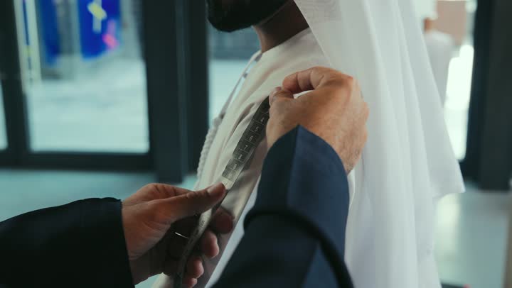 Skill in mastering youth clothing tailoring, men's clothing tailoring and tailoring shop, concept of the world of fashion and style, mastery of making clothes, practicing the tailoring profession, close-up of a young Emirati Gulf man wearing a navy blue kandura taking measurements of a customer's body using a tape measure, handicrafts and crafts.