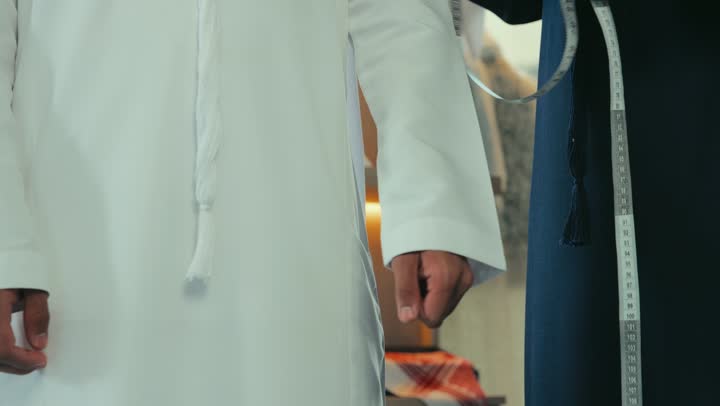 The concept of the world of fashion and style, mastery of making clothes, practicing the tailoring profession, the skill of mastering the detailing of youth clothes, a men's tailoring and detailing shop, a close-up of a young Emirati Gulf man wearing a navy blue kandura taking measurements of the customer's body using a measuring tape, manual crafts and professions.