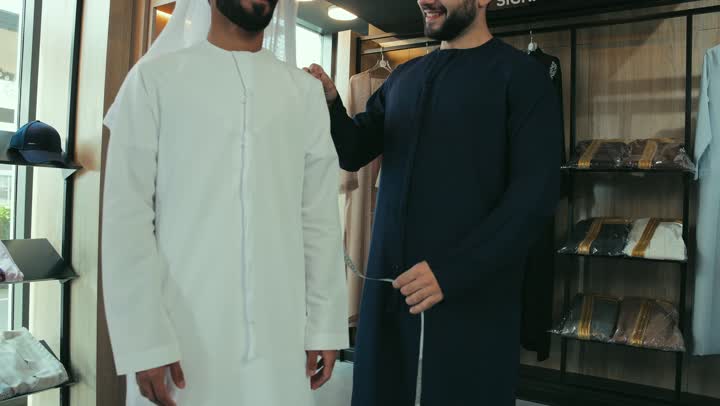 Mastering clothing, practicing the tailoring profession, skill of mastering tailoring youth clothes, men's tailoring and tailoring shop, close-up of a young Emirati Gulf man wearing a navy blue kandura taking measurements of a customer's body using a tape measure, concept of the world of fashion and style, handicrafts and crafts.
