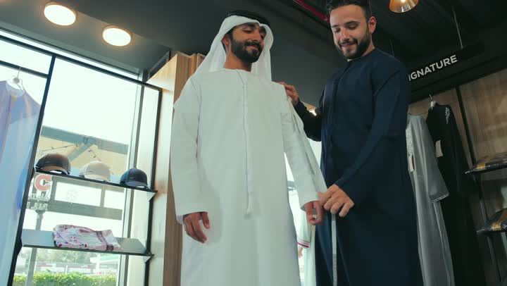 ممارسة مهنة الخياطة، مهارة اتقان تفصيل الملابس الشبابية، محل خياطة و تفصيل الملابس الرجالي ، تصوير مقرب لشاب خليجي اماراتي يرتدي الكندورة الكحلي يقوم بأخذ القياسات الخاصة بجسم الزبون باستخدام الشريط المتري ، اتقان صنع الملابس، مفهوم عالم الازياء والموضه ، حرف ومهن يدوية .